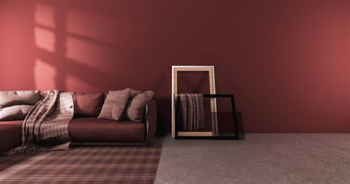 A cozy living room with a maroon wall, a modern couch adorned with pillows and a blanket, and framed textiles leaning against the wall.