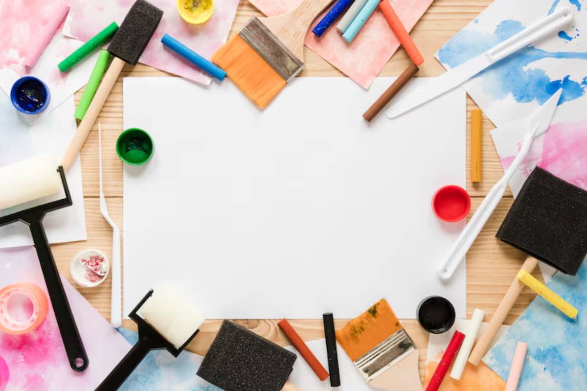 Art supplies are spread around a blank canvas, featuring brushes, rollers, paints, and colorful papers on a wooden surface.