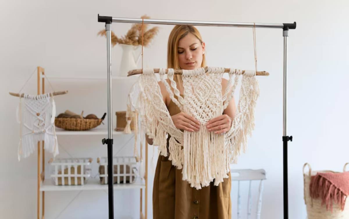 A person holds a large, intricate macramé wall hanging, surrounded by cozy home decor items in a bright, minimalist setting.