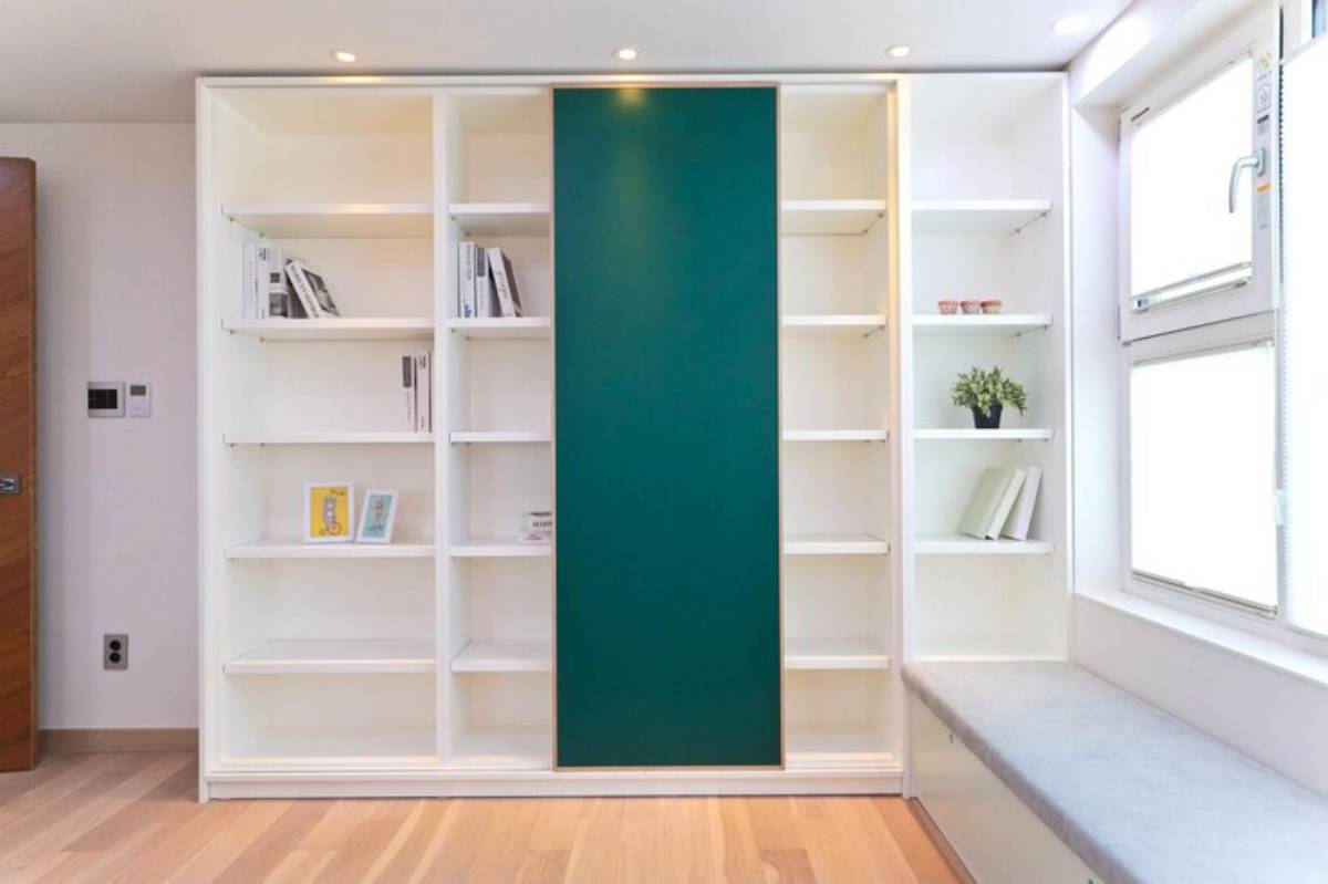 Modern white bookshelf with a teal sliding panel, books, and decor accents, beside a window and cushioned bench.