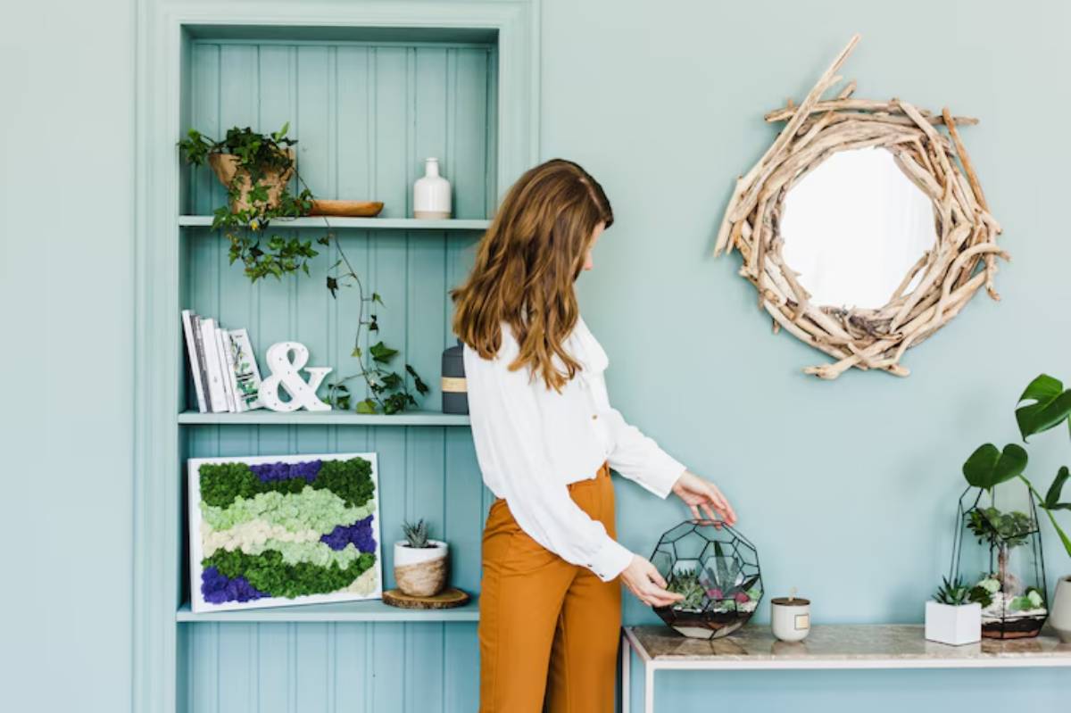 A woman in a white blouse and orange pants arranges a geometric terrarium on a table, surrounded by greenery and decor on a pastel wall.