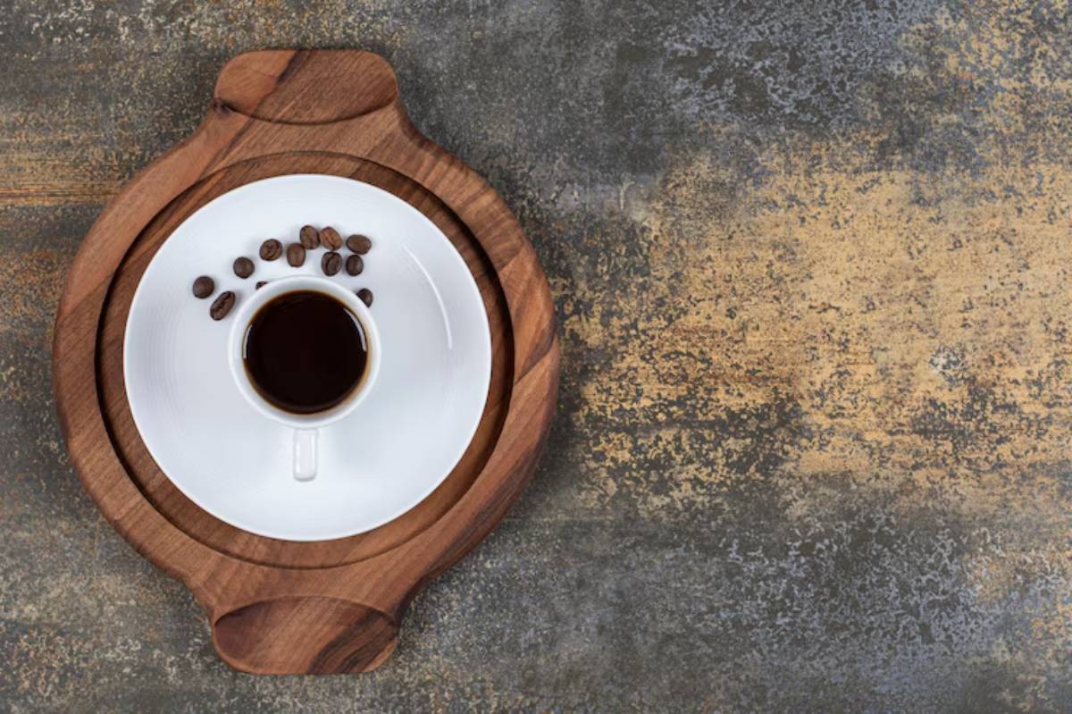 A white coffee cup on a round wooden coaster, accompanied by scattered coffee beans on a textured stone surface.