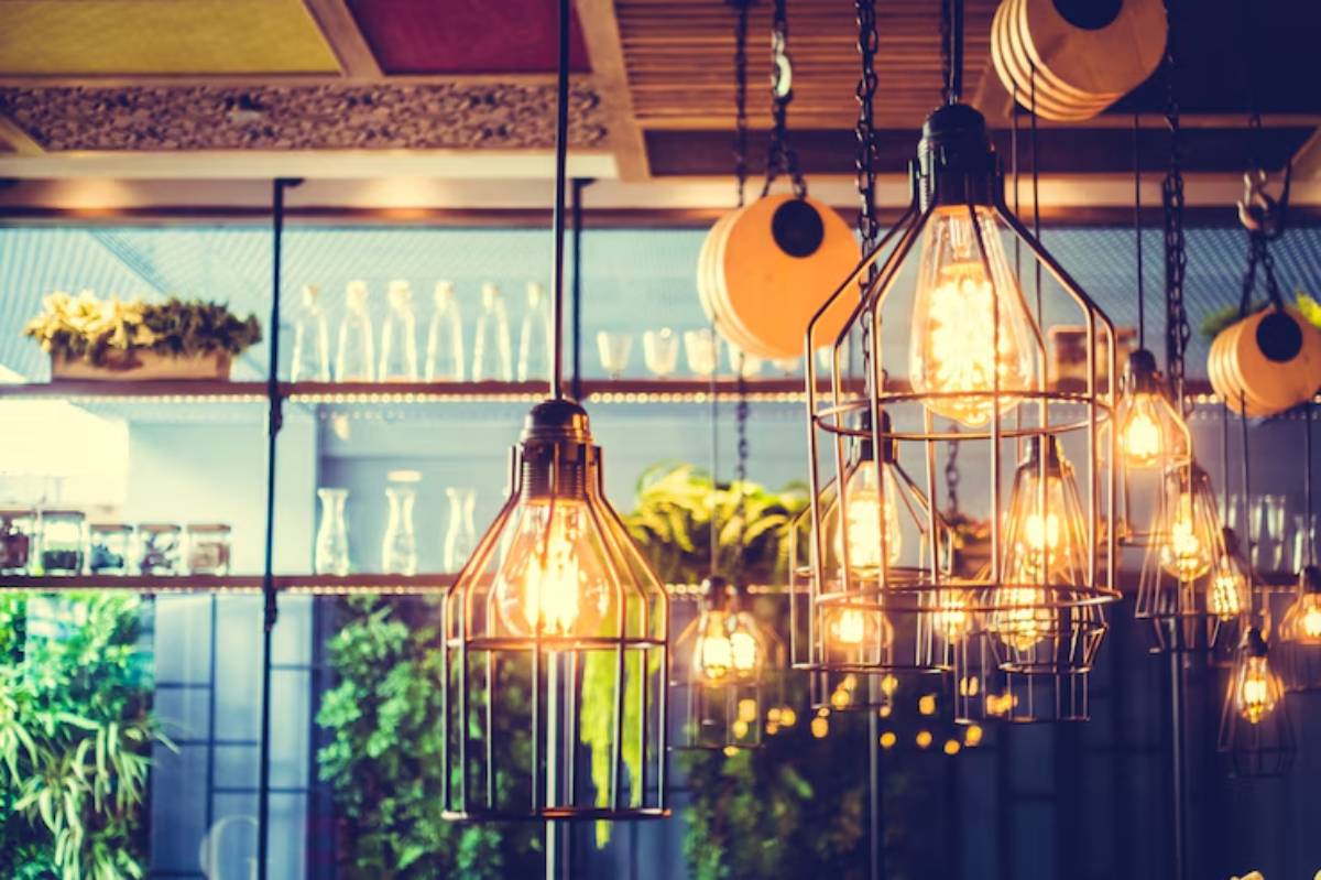 A cozy restaurant interior featuring various hanging light fixtures and a backdrop of greenery and glassware, creating a warm ambiance.