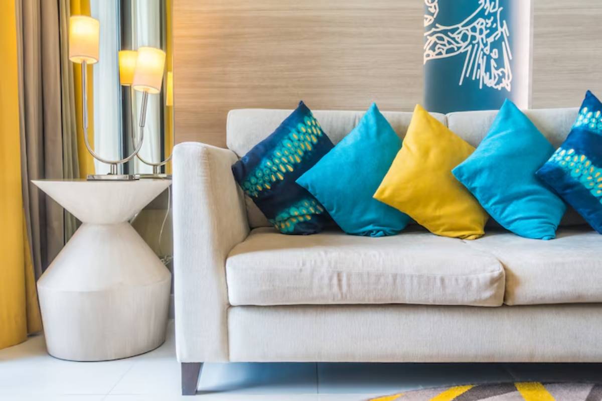 A cozy sofa adorned with vibrant blue and yellow cushions next to a modern lamp and side table, set against warm, neutral tones.