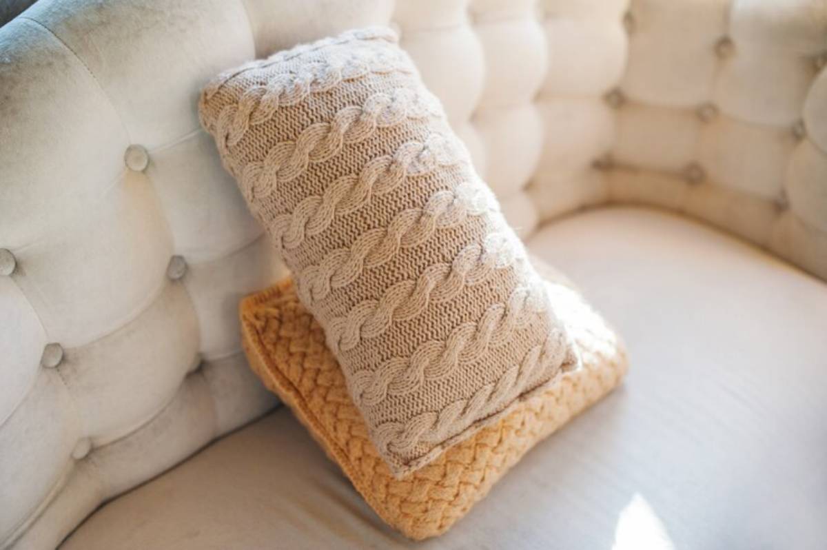 A textured beige knitted pillow with a cable design rests on a cozy yellow pillow, both placed on a tufted light-colored couch.
