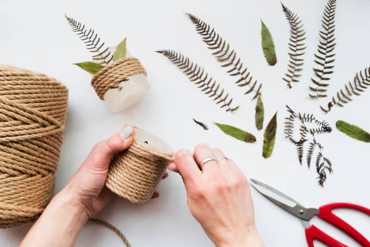 A hand wraps twine around a glass pot with ferns, surrounded by additional ferns and scissors on a white surface.