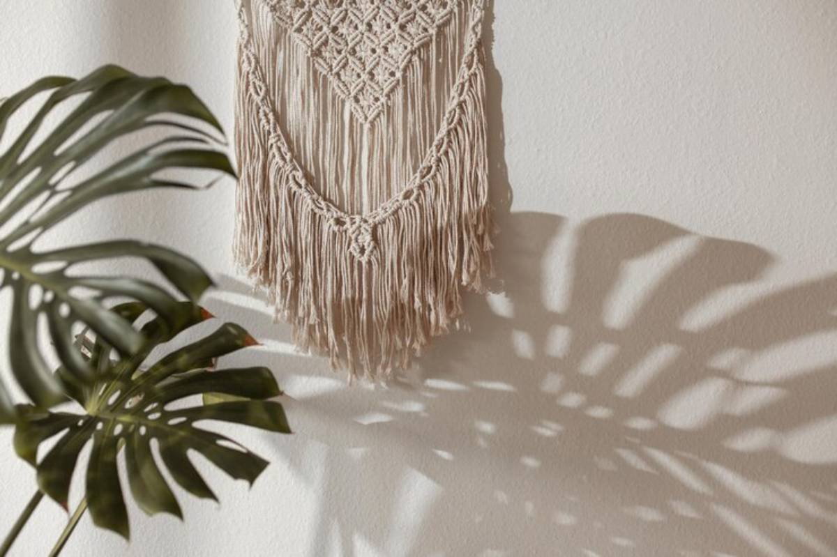 A woven macramé wall hanging casts intricate shadows, accompanied by a vibrant green monstera leaf in soft lighting.