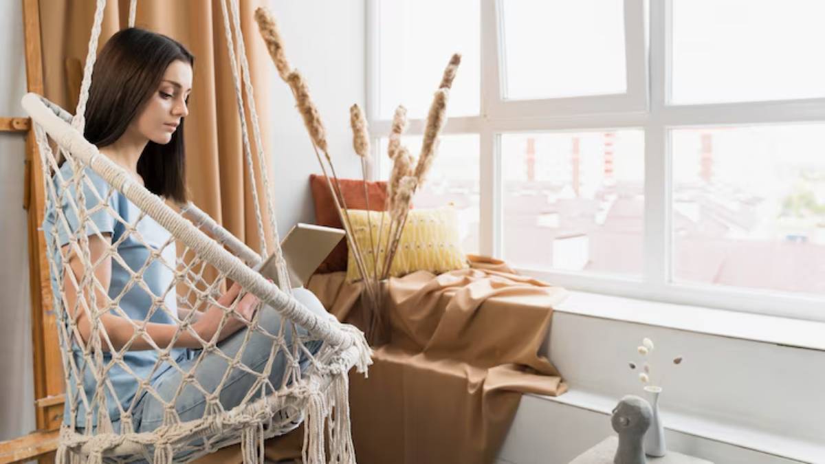 A person sits on a macrame swing by a bright window, surrounded by decorative plants and cozy fabrics.