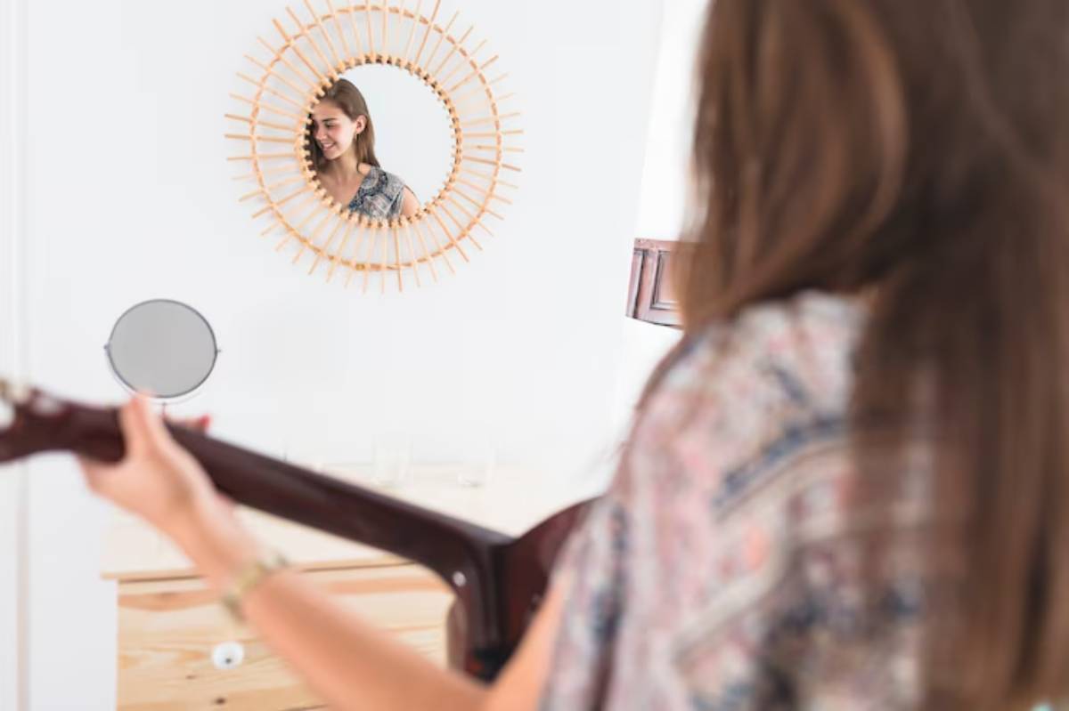A person holding a guitar stands in front of a decorative round mirror on a light wall, with soft natural lighting in a cozy room.