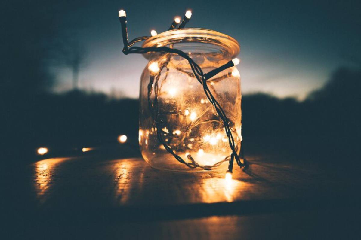 A illuminated jar filled with string lights rests on a wooden surface, set against a twilight sky. Soft glow enhances the tranquil scene.