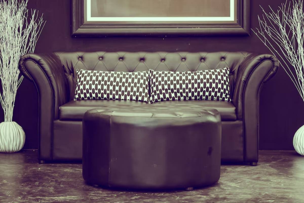 A stylish black leather sofa with tufted design and decorative patterned cushions, accompanied by a round ottoman and minimalist decor.