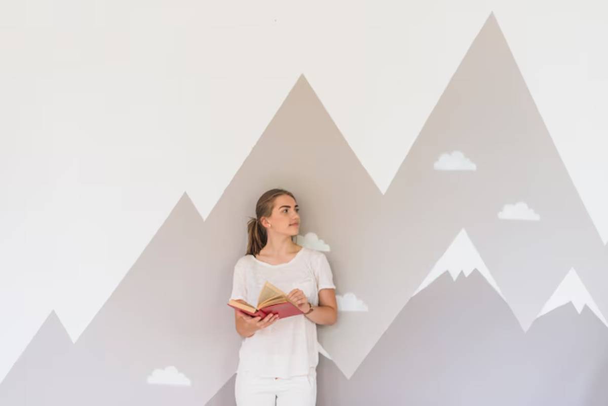 A person in a white shirt holds a book against a minimalist wall featuring painted gray mountains and white clouds.