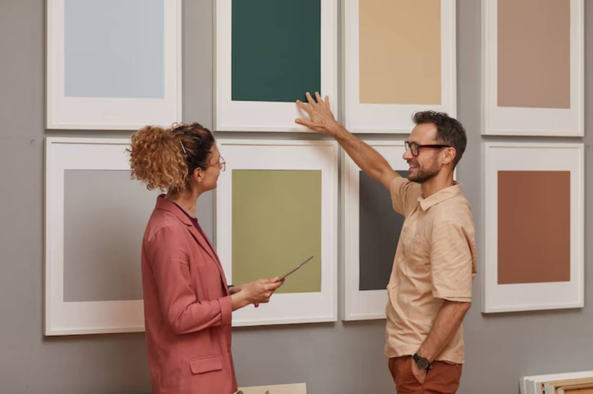 A woman in a pink blazer and a man in a beige shirt discuss color samples displayed on a wall, evaluating options together.