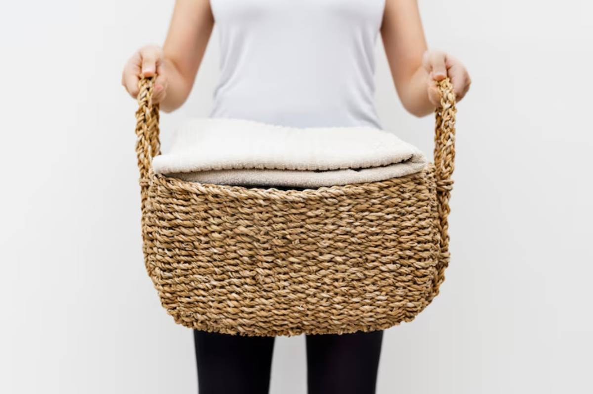 A person holding a woven basket filled with folded blankets, standing against a plain white background, dressed in a casual top.
