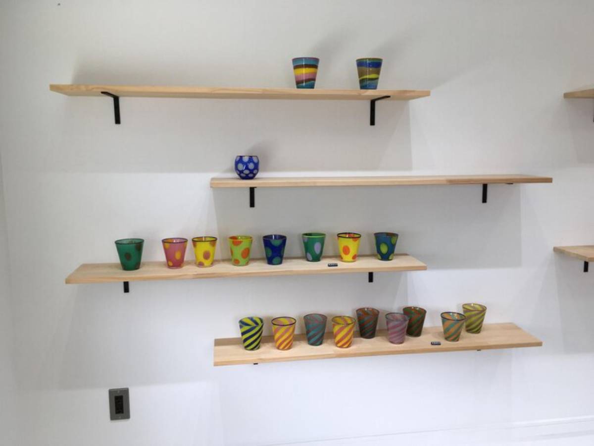Colorful patterned glass cups displayed on wooden shelves against a white wall, showcasing vibrant designs and hues.