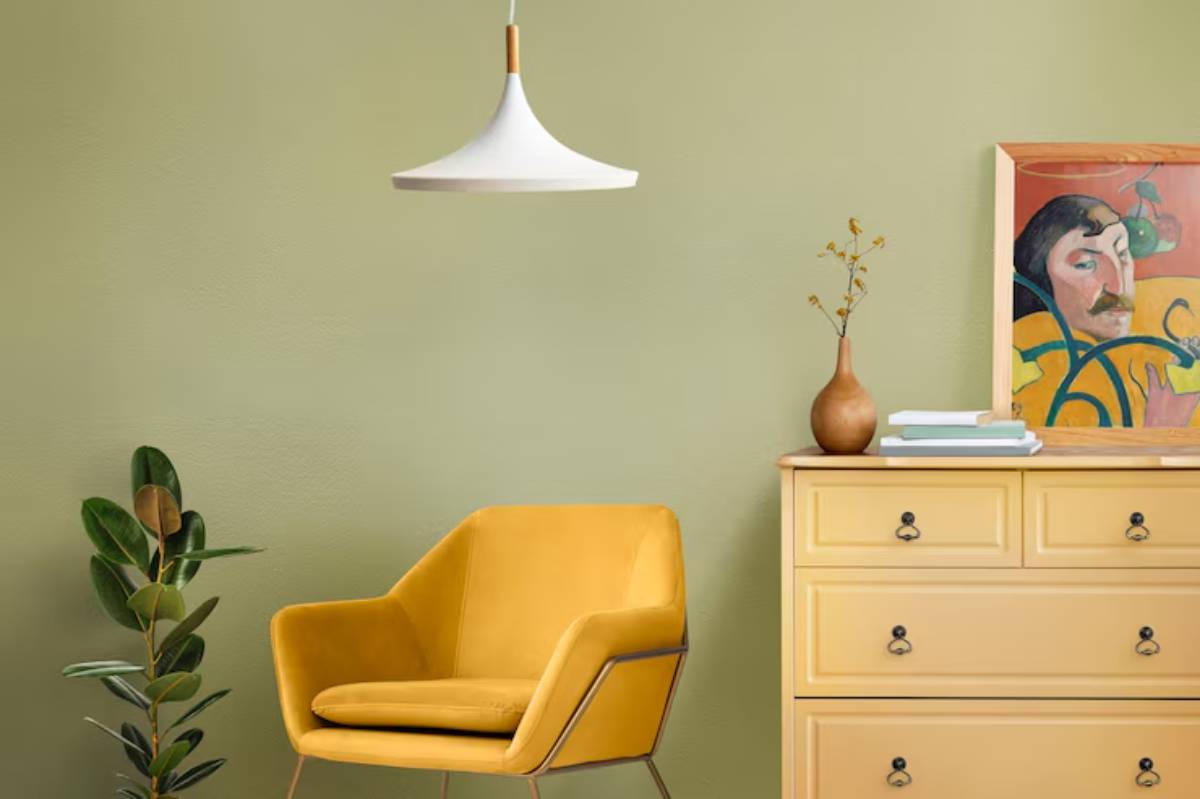 Cozy interior featuring a yellow armchair next to a wooden dresser, potted plant, and a colorful framed artwork against a green wall.