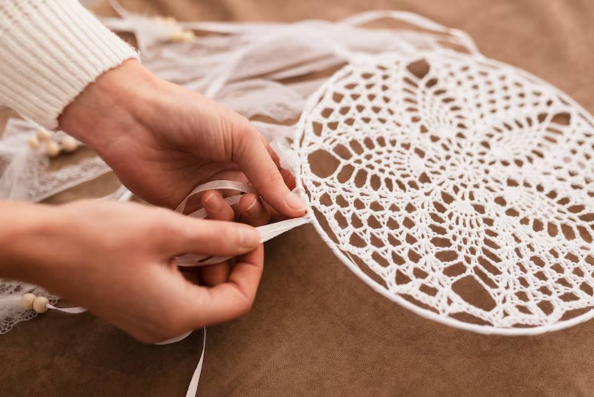A person's hands delicately tie ribbons onto a beautiful, intricate white lace dreamcatcher on a brown surface.
