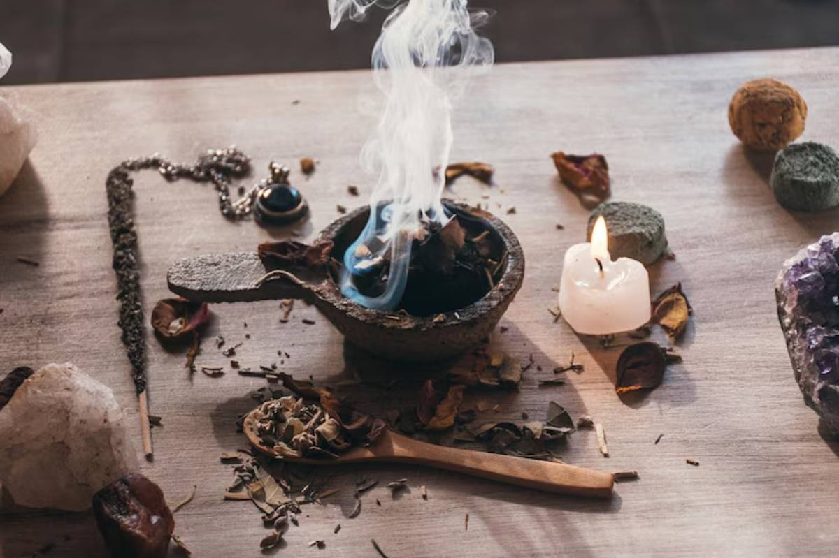 A mortar filled with smoke from burning herbs, surrounded by candles, stones, and dried leaves on a wooden surface.