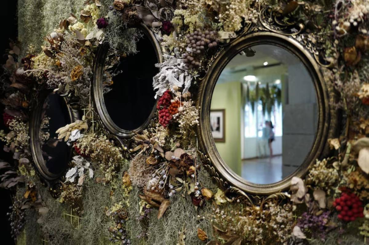 Three ornate, floral-framed mirrors adorned with dried flowers and moss, reflecting a softly lit gallery space behind.