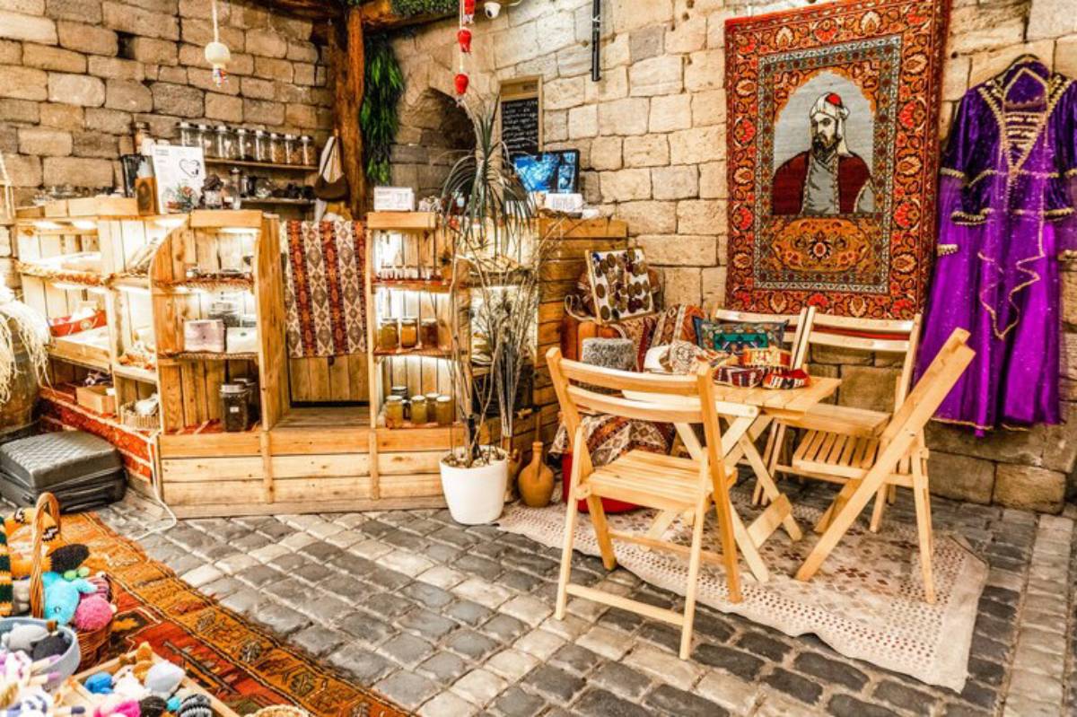 A cozy shop interior featuring wooden shelving, decorative rugs, and colorful cushions, with a rustic stone wall backdrop.