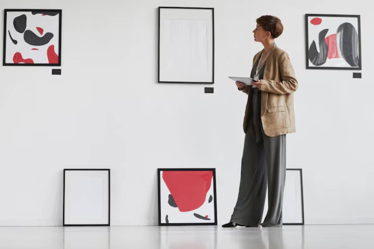 A person stands in an art gallery, observing abstract paintings on a white wall, with additional frames placed on the floor.