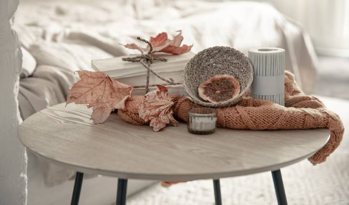 A cozy table setting with dried leaves, a white candle, a knitted scarf, and neatly stacked books, exuding autumn warmth.