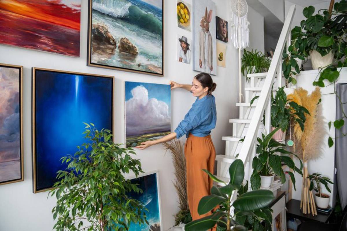 A woman in a blue shirt and orange pants arranges colorful paintings on a white wall adorned with plants and decorative items.