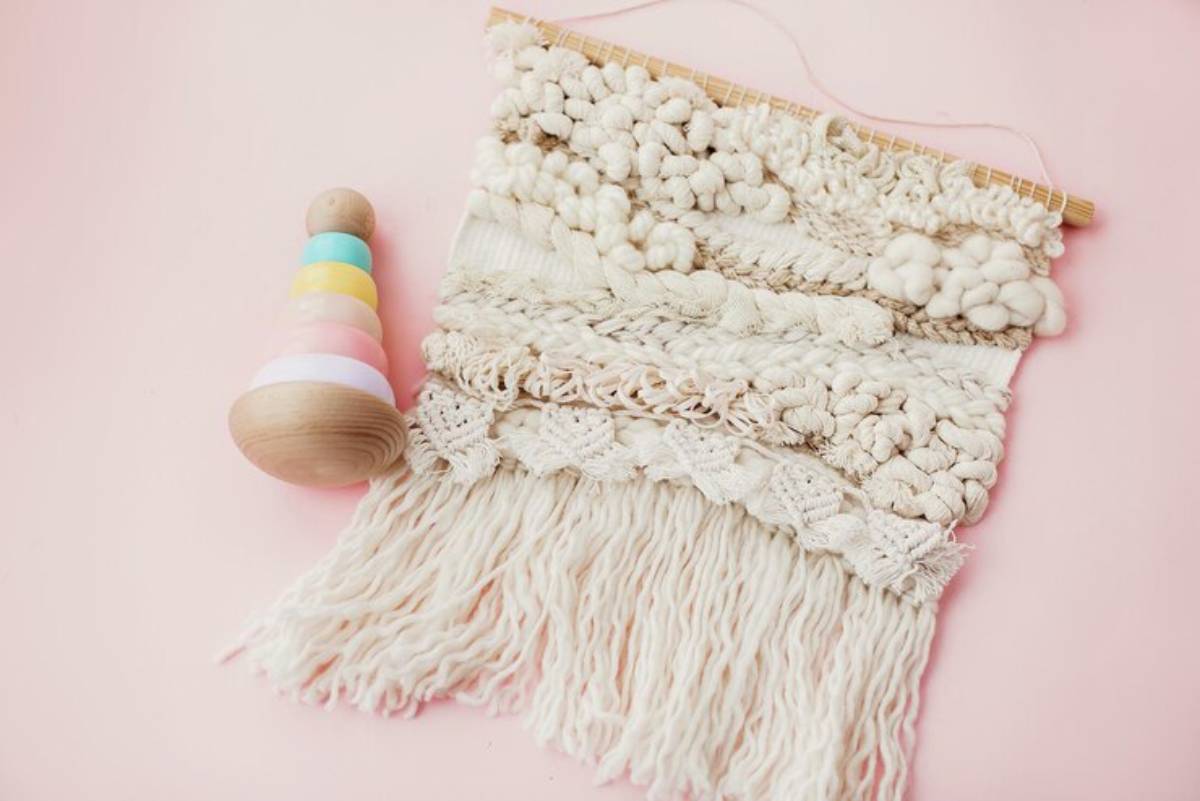 A colorful wooden stacking toy beside a textured white macramé wall hanging on a pink background.
