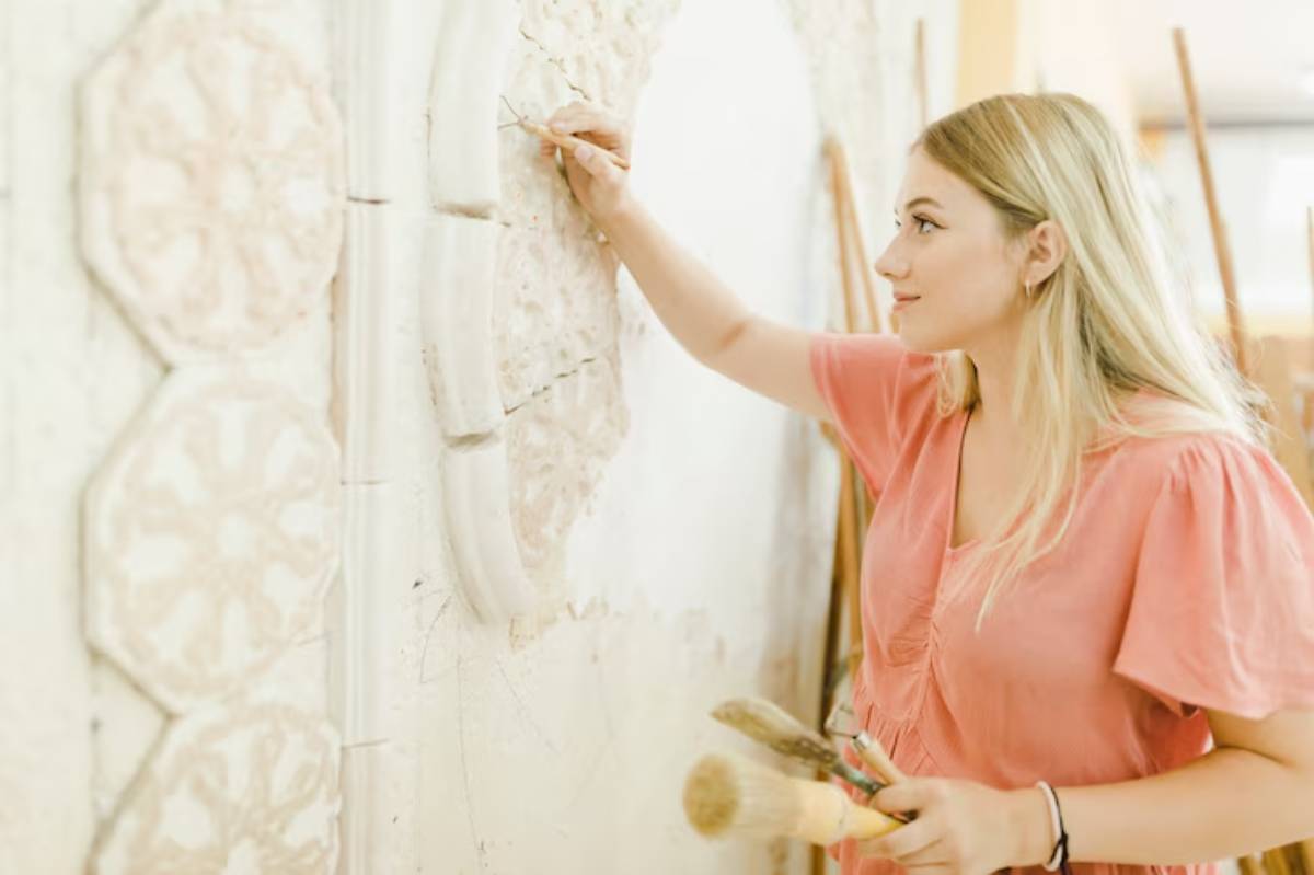 A young woman in a pink shirt is intricately carving a decorative wall piece, surrounded by art tools and wooden sticks.