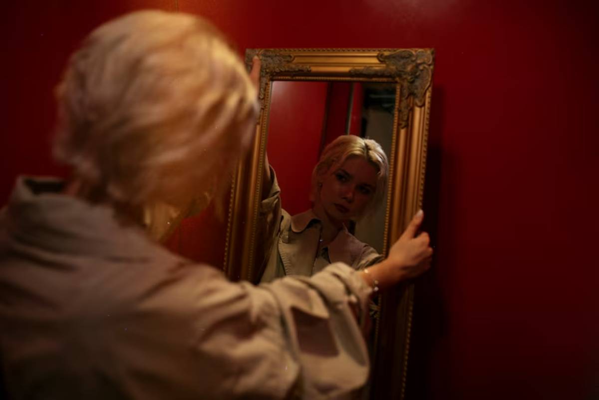 A person with blonde hair in a light outfit gazes into an ornate mirror against a deep red wall, creating a reflective moment.