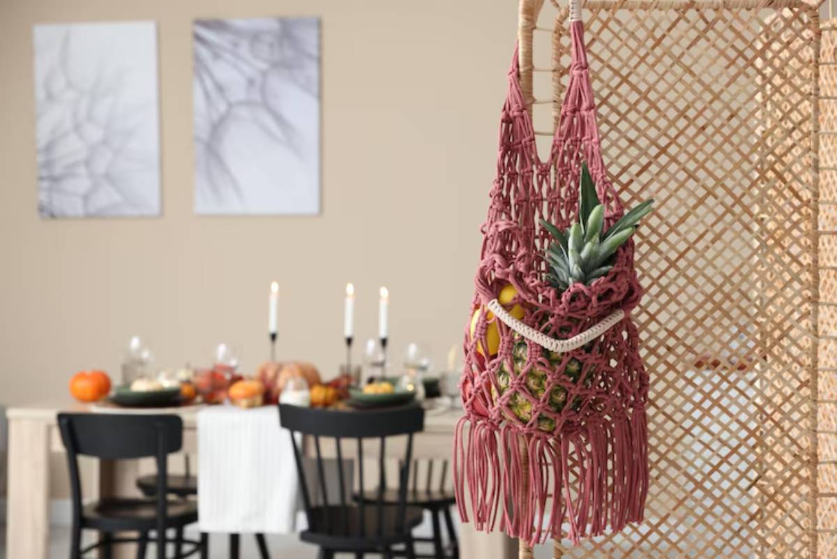 A pink macramé bag filled with fruits hangs against a woven backdrop, overlooking a cozy dining table set for a gathering.