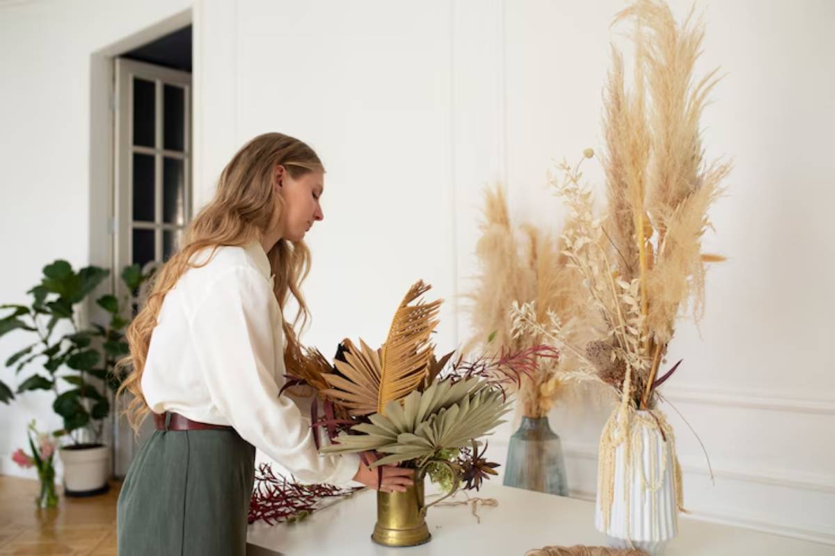A person arranges dried plants in a gold vase on a white table, surrounded by other elegant floral arrangements in a bright room.