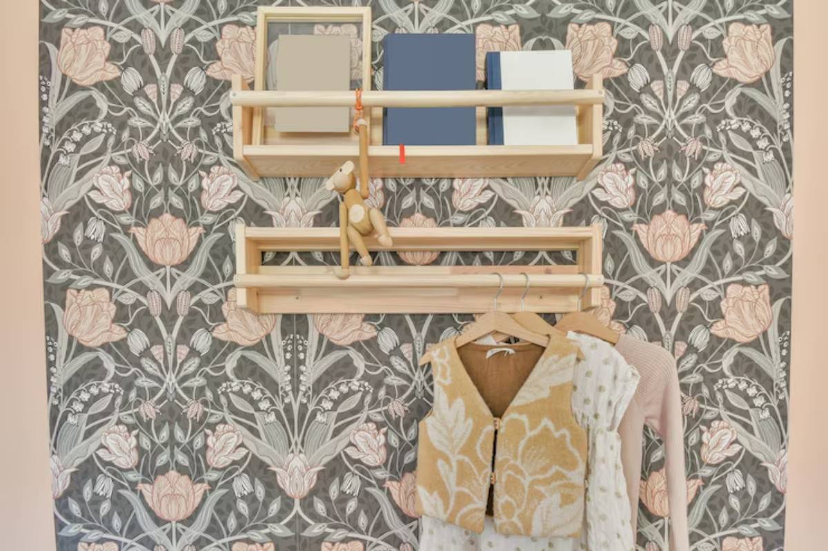 A stylish wall display featuring light wood shelves with books, a toy monkey, and hanging children's clothing against a floral wallpaper.