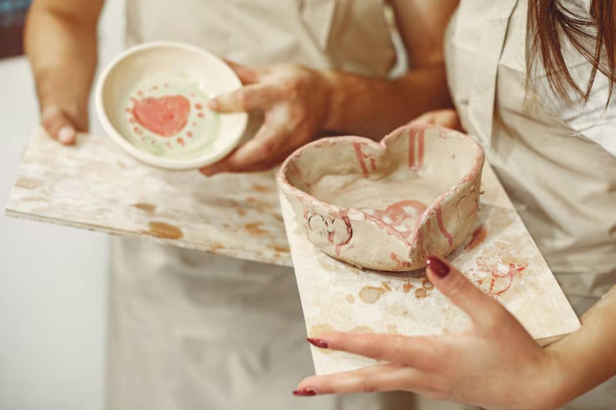 Two hands hold handmade pottery: a heart-shaped bowl and a shallow dish with a painted heart design, on a wooden surface.