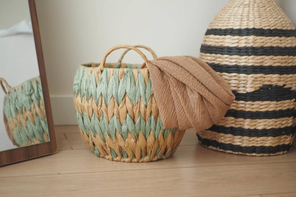 A woven basket with mint green and natural straw patterns holds a brown fabric, beside a tall striped straw basket and a mirror.