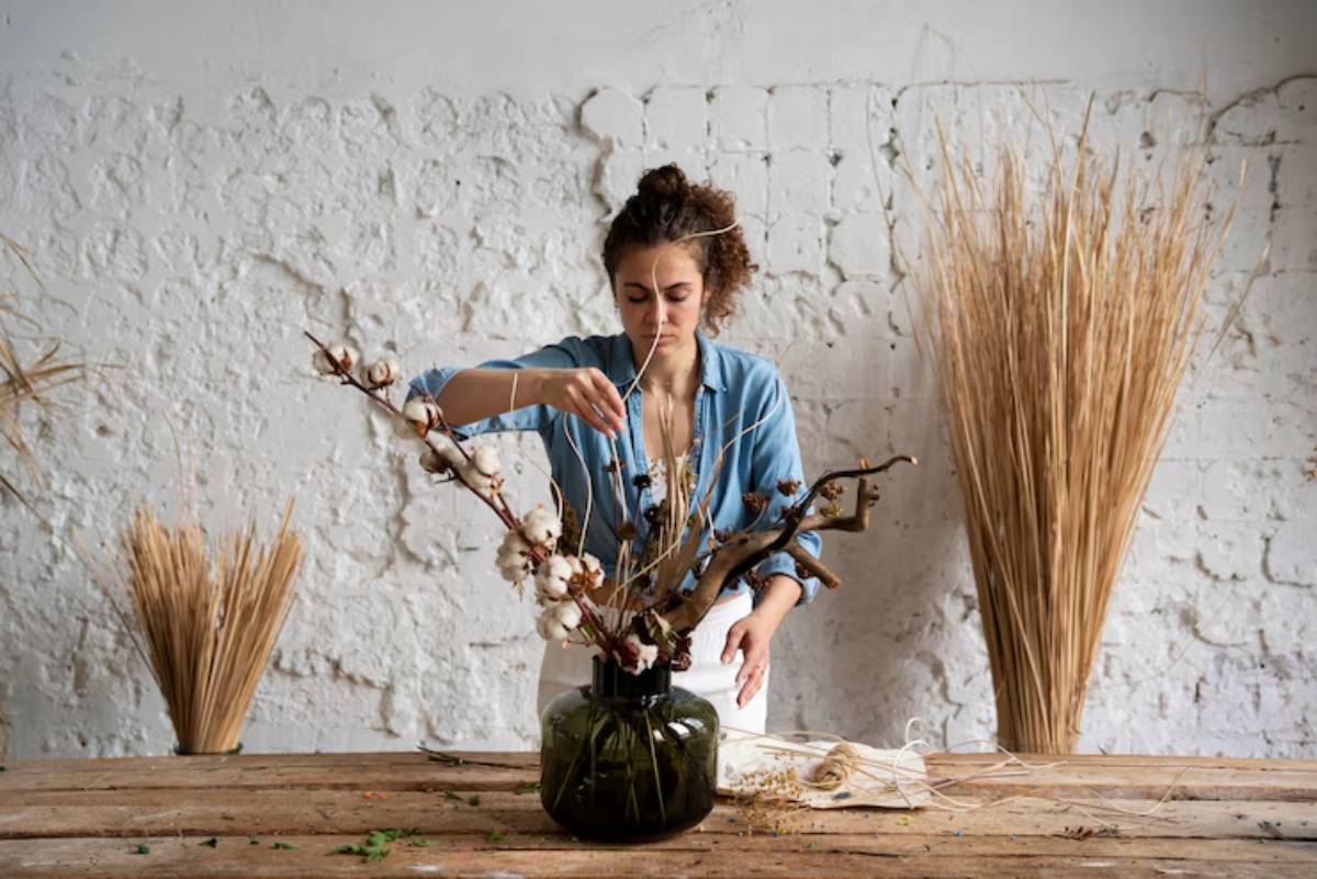 A person arranges cotton branches and dried flowers in a dark glass vase on a wooden table, surrounded by decorative grasses.