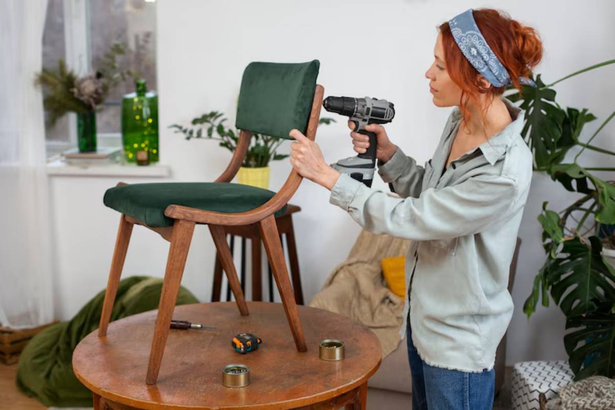 A person uses a power drill to assemble a green cushioned chair on a wooden table surrounded by plants and tools.