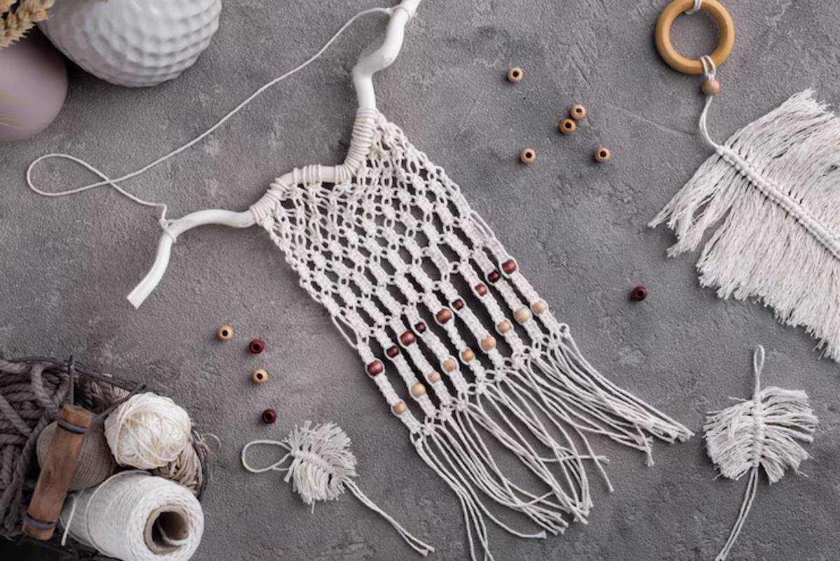 A white macramé wall hanging with wooden beads and fringe lies on a grey surface, surrounded by crafting materials.