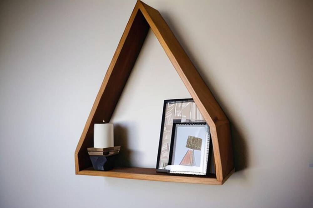 A triangular wooden shelf on a wall displays a white candle, framed artwork, and a decorative piece.